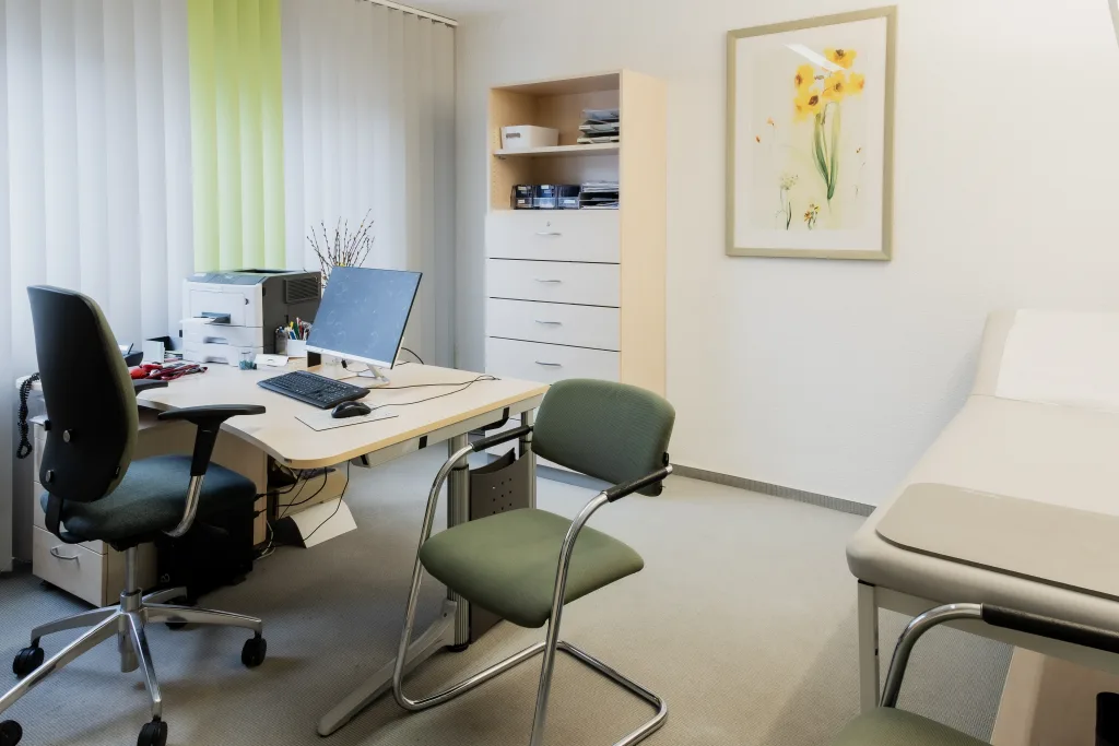 Foto von einem leeren Behandlungsraum mit Fenster, in dem links ein Schreibtisch mit Computer steht. Rechts im Raum befindet sich eine Behandlungsliege. An der hinteren Wand hängt ein großes Bild einer Blume. 